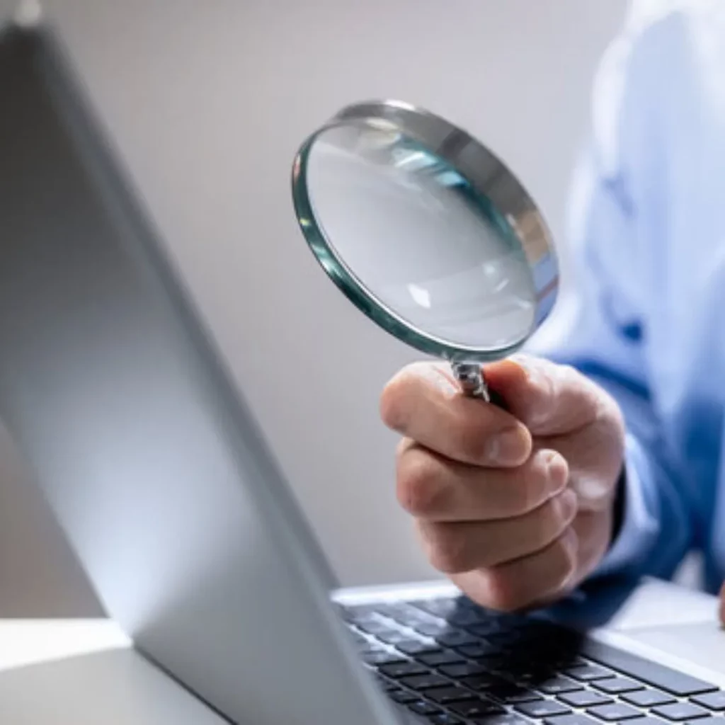 A man closely examines a laptop screen with a magnifying glass.