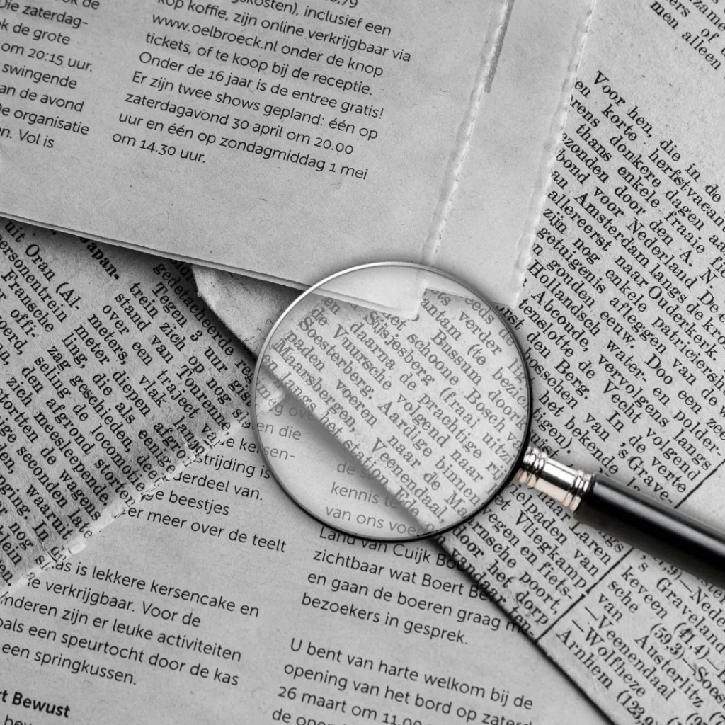 A man with short brown hair and glasses holds a magnifying glass in his right hand. He is closely examining a laptop screen that displays a complex diagram with lines, shapes, and text in multiple languages, including what appears to be Dutch.