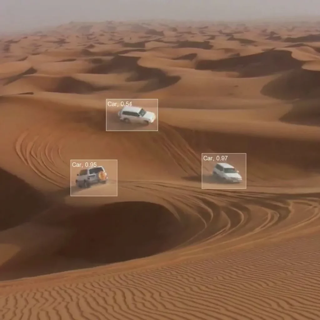 The image shows two cars driving through a desert landscape. Text on the image reads "Car, 0.54", "Car, 0.95" and "Car, 0.97" which likely refers to the confidence level of an object detection model.